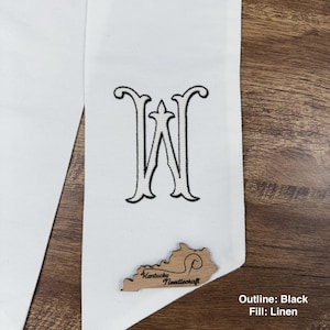 May include: White graduation stole with a black embroidered letter "W" and a wooden Kentucky-shaped embellishment. The text "Kentucky Needlecraft" is visible on the wooden embellishment. The text "Outline: Black Fill: Linen" is also visible.