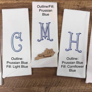 May include: Five white monogrammed hand towels with blue embroidered letters. The letters are outlined and filled with different shades of blue. The towels are folded and displayed on a wooden surface.
