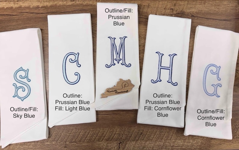 May include: Five white monogrammed hand towels with blue embroidered letters. The letters are outlined and filled with different shades of blue. The towels are folded and displayed on a wooden surface.