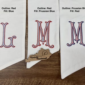 May include: Three white fabric banners with embroidered letters. The first banner has the letter "L" with a red outline and blue fill. The second banner has the letter "M" with a red outline and Prussian blue fill. The third banner has the letter "M" with a Prussian blue outline and red fill.