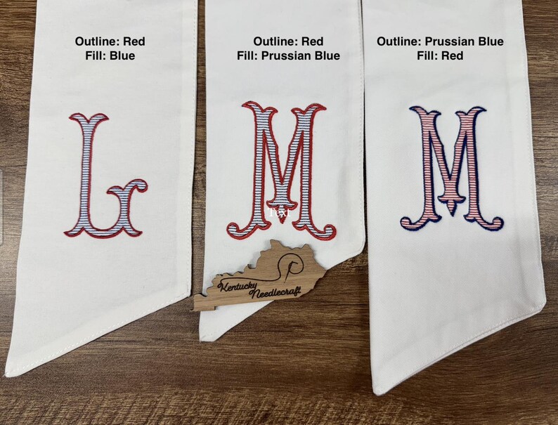 May include: Three white fabric banners with embroidered letters. The first banner has the letter "L" with a red outline and blue fill. The second banner has the letter "M" with a red outline and Prussian blue fill. The third banner has the letter "M" with a Prussian blue outline and red fill.