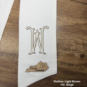 May include: A white sash with a large embroidered letter "W" in light brown with a beige fill. A wooden cutout of the state of Kentucky with the text "Kentucky Needlecraft" is attached to the sash. The text "Outline: Light Brown Fill: Beige" is visible.