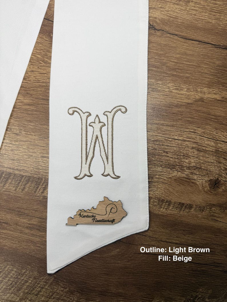 May include: A white sash with a large embroidered letter "W" in light brown with a beige fill. A wooden cutout of the state of Kentucky with the text "Kentucky Needlecraft" is attached to the sash. The text "Outline: Light Brown Fill: Beige" is visible.