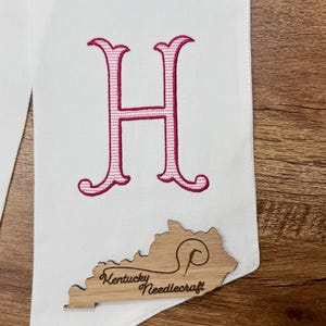 May include: White fabric with a large, embroidered pink letter "H". A wooden cutout of Kentucky with "Kentucky Needlecraft" is attached. Text at the bottom reads "Outline: Dark Fuschia Fill: Pink".