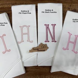 May include: Four white monogrammed hand towels with different letters embroidered in pink. The text on the towels reads "Outline & Fill: Flesh Pink", "Outline & Fill: Dark Fuschia", "Outline: Salmon Pink Fill: Flesh Pink", and "Outline & Fill: Pink". A wooden Kentucky Needlecraft cutout is also present.