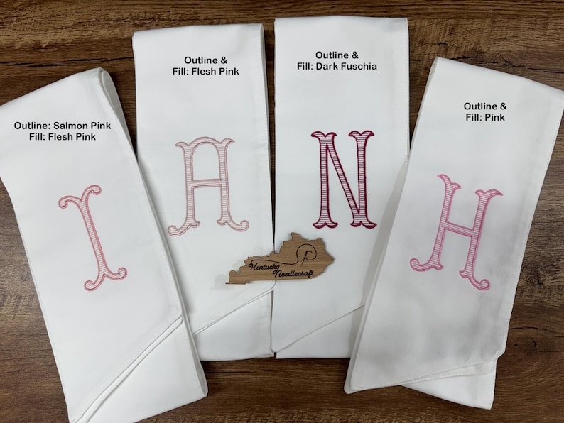 May include: Four white monogrammed hand towels with different letters embroidered in pink. The text on the towels reads "Outline & Fill: Flesh Pink", "Outline & Fill: Dark Fuschia", "Outline: Salmon Pink Fill: Flesh Pink", and "Outline & Fill: Pink". A wooden Kentucky Needlecraft cutout is also present.