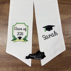 May include: White graduation stole with embroidered details. Features "Class of 2026" in a green and gold shield design, a graduation cap, and the name "Sarah". Includes a Kentucky Needlecraft logo.