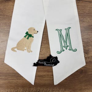May include: White graduation sash with embroidered details. Features a tan dog with a green bow, a large green letter "M", and a black Kentucky outline with "Kentucky Needlecraft" text.