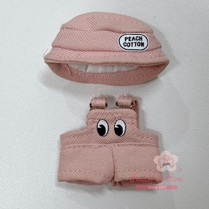 May include: A light pink doll-sized outfit set, including a bucket hat and overalls. The hat has a "PEACH COTTON" patch. The overalls feature cartoon eyes. The set is made of a textured fabric.