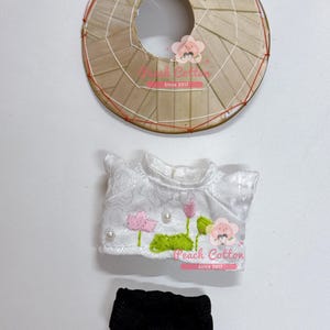 以下が含まれることがあります： 円錐形の帽子、白いトップス、花の刺繍、黒いショートパンツを含む、ミニチュアの伝統的なベトナムの衣装。帽子は編まれたわらと赤い糸でできています。トップスにはピンクの蓮の花と緑の葉が描かれています。