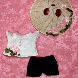 May include: A doll-sized outfit set, including a white floral jacket with pink lotus flower accents, black shorts, and a decorative hat. The hat is beige with pink and green floral details. The background is a pink floral pattern. Text reads "Handmade by Peach Cotton."