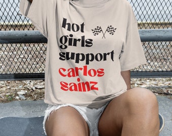 May include: A beige t-shirt with the text "hot girls support carlos sainz" in black and red lettering. The text is in a bold, retro font. Two black and white chequered flags are crossed above the text.