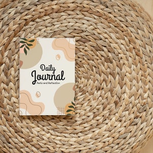 May include: A white journal with the text "Daily Journal" and "Parts and Reflection" on the cover. The cover has a brown, peach, and green abstract design. The journal is sitting on a woven straw mat.