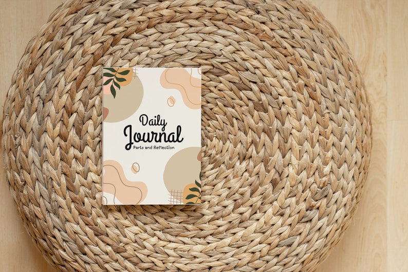 May include: A white journal with the text "Daily Journal" and "Parts and Reflection" on the cover. The cover has a brown, peach, and green abstract design. The journal is sitting on a woven straw mat.
