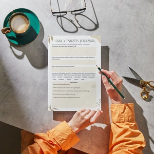 May include: A person is filling out a "Daily Parts Journal" worksheet. The worksheet asks questions about how the person is feeling and what parts of themself they are focusing on. The worksheet is on a gray surface with a cup of coffee and a pair of scissors.
