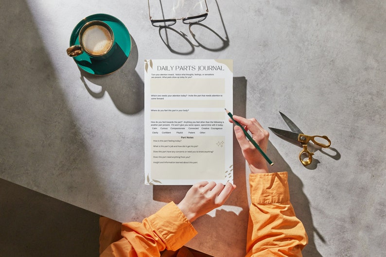 May include: A person is filling out a "Daily Parts Journal" worksheet. The worksheet asks questions about how the person is feeling and what parts of themself they are focusing on. The worksheet is on a gray surface with a cup of coffee and a pair of scissors.