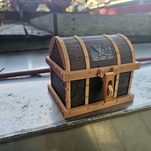 Puede incluir: Un peque&ntilde;o cofre del tesoro de madera con un acabado marr&oacute;n oscuro y adornos marrones claros. El cofre presenta detalles met&aacute;licos y un cierre de pestillo. La parte superior tiene una placa met&aacute;lica decorativa. Un art&iacute;culo decorativo.