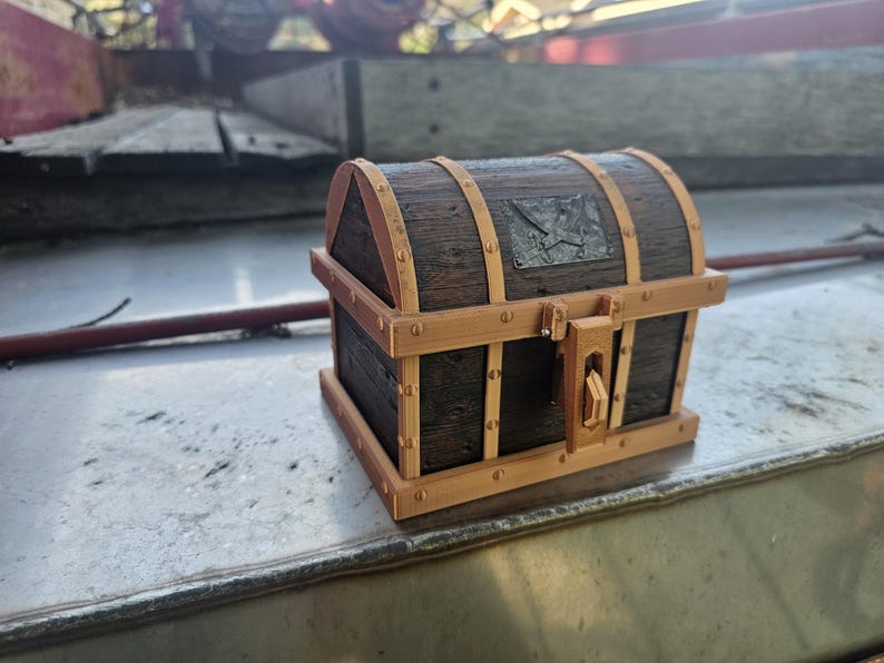 Puede incluir: Un peque&ntilde;o cofre del tesoro de madera con un acabado marr&oacute;n oscuro y adornos marrones claros. El cofre presenta detalles met&aacute;licos y un cierre de pestillo. La parte superior tiene una placa met&aacute;lica decorativa. Un art&iacute;culo decorativo.
