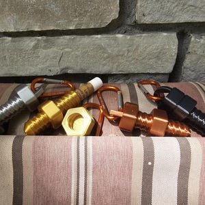 Op de afbeelding: Een verzameling metalen sleutelhangers in de vorm van bouten in zilver, goud, koper en zwart. Elke sleutelhanger heeft een karabijnhaak. De sleutelhangers liggen op een gestreept bruin en beige kussen, met een stenen muur op de achtergrond.