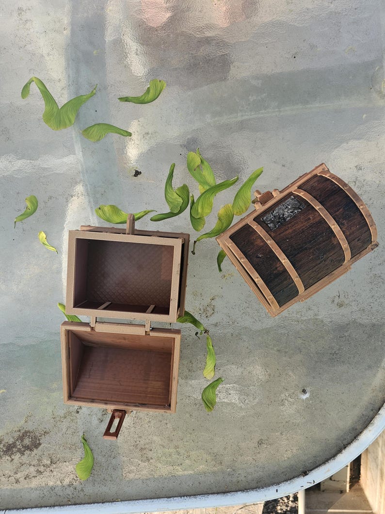 Puede incluir: Dos peque&ntilde;os cofres de madera, uno abierto y otro cerrado, con acabado marr&oacute;n oscuro y ribete marr&oacute;n claro. Semillas verdes de arce esparcidas alrededor de los cofres sobre una superficie de vidrio.