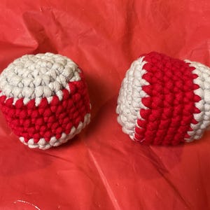 Puede incluir: Dos pelotas redondas tejidas a ganchillo con una superficie texturizada. Cada pelota está hecha con hilo gris claro y una franja vertical ancha y prominente de hilo rojo brillante en el centro, imitando el aspecto de una pelota de béisbol.