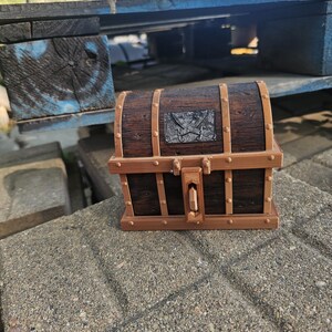 Puede incluir: Un peque&ntilde;o cofre del tesoro de madera con un acabado marr&oacute;n oscuro y detalles en color bronce. El cofre presenta una placa met&aacute;lica decorativa con un dise&ntilde;o de espadas cruzadas. El cofre est&aacute; sobre una superficie de hormig&oacute;n.