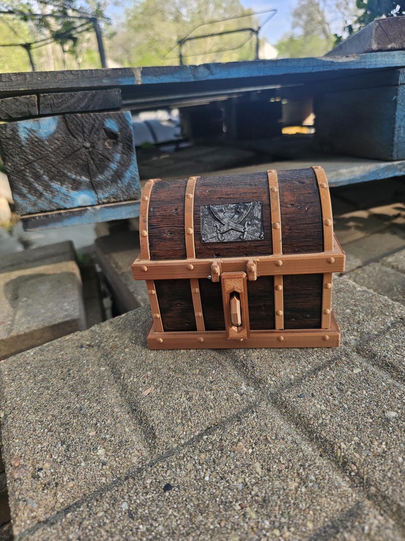 Puede incluir: Un peque&ntilde;o cofre del tesoro de madera con un acabado marr&oacute;n oscuro y detalles en color bronce. El cofre presenta una placa met&aacute;lica decorativa con un dise&ntilde;o de espadas cruzadas. El cofre est&aacute; sobre una superficie de hormig&oacute;n.