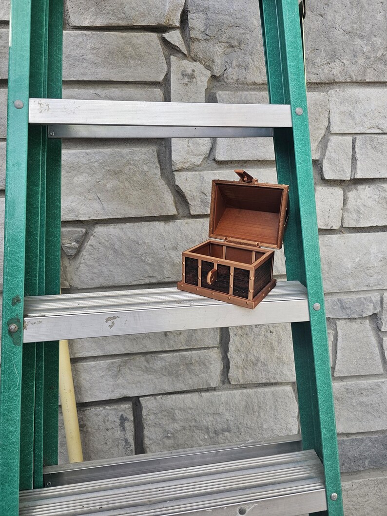 Puede incluir: Un peque&ntilde;o cofre del tesoro de madera abierto, con detalles en marr&oacute;n oscuro y negro, descansa sobre un pelda&ntilde;o de escalera met&aacute;lica. El cofre tiene un cierre color lat&oacute;n y est&aacute; colocado contra una pared de piedra gris. La escalera es verde.