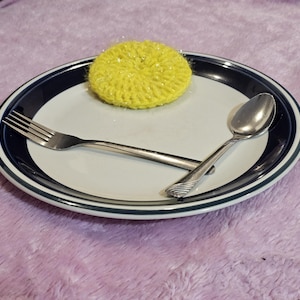 May include: A white plate with a navy blue rim, a silver fork and spoon, and a yellow crocheted item. The fork and spoon are crossed on the plate. The crocheted item is round and textured.