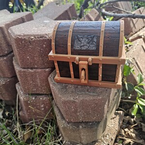 Puede incluir: Un peque&ntilde;o cofre del tesoro de madera, con acabado marr&oacute;n oscuro y detalles en marr&oacute;n claro. El cofre presenta un emblema met&aacute;lico con espadas cruzadas y una cerradura. Est&aacute; colocado sobre una pila de ladrillos rojos.