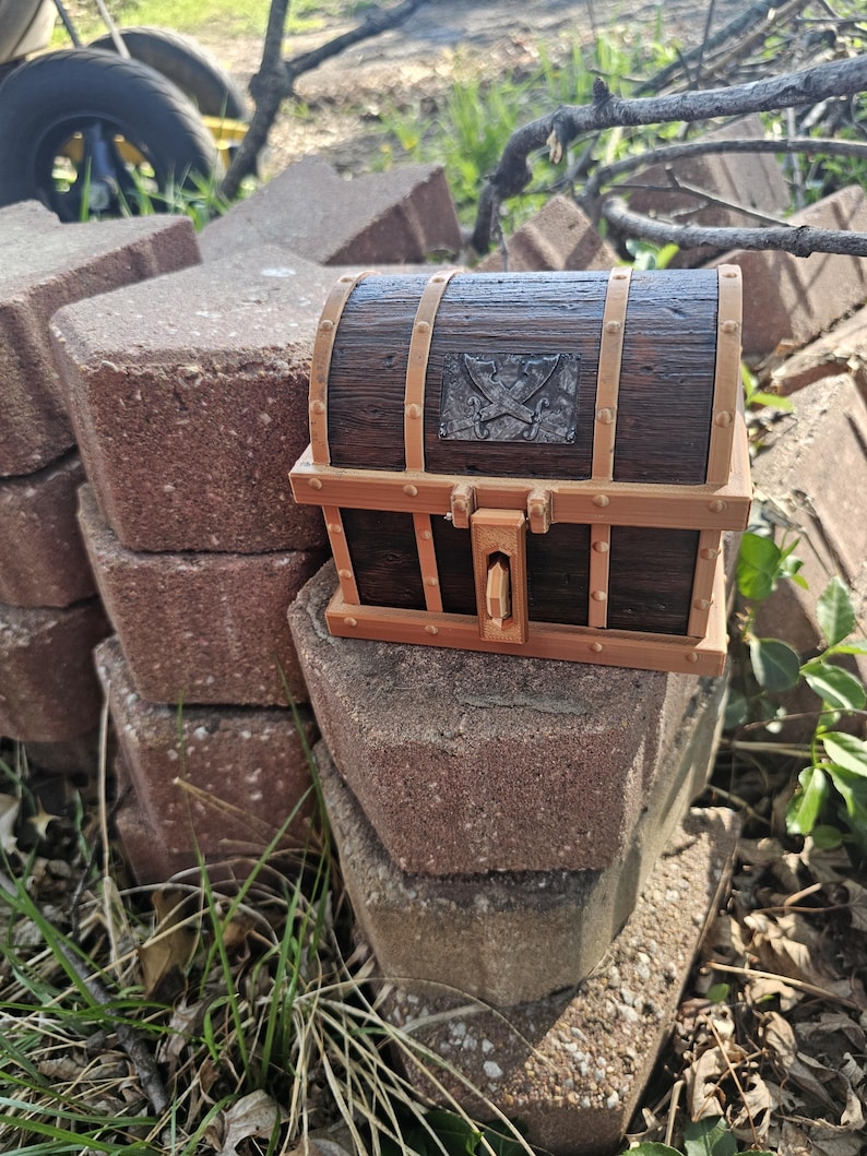 Puede incluir: Un peque&ntilde;o cofre del tesoro de madera, con acabado marr&oacute;n oscuro y detalles en marr&oacute;n claro. El cofre presenta un emblema met&aacute;lico con espadas cruzadas y una cerradura. Est&aacute; colocado sobre una pila de ladrillos rojos.