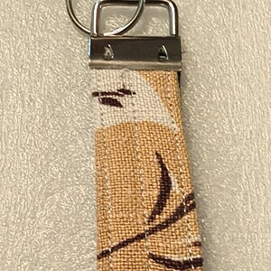 May include: A fabric keychain featuring a tan and white textured material with a dark brown abstract leaf pattern. The fabric is secured by a silver-toned metal clamp and includes a circular silver-toned metal split ring for attaching keys.