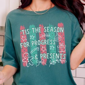 May include: A dark teal short-sleeve t-shirt featuring a graphic print with the text 'TIS THE SEASON FOR PROGRESS & PRESENTS' in white. The design includes red and green vertical brush strokes, a gingerbread man, a gift box, and stars.
