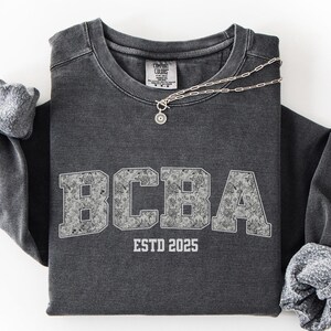 May include: A dark grey sweatshirt featuring the letters "BCBA" in a textured, light grey font. Below the letters is "ESTD 2025". The sleeves are cuffed. A silver necklace with a pendant is draped on the shirt. The word "PEPPER" is in a teal box in the bottom left corner.