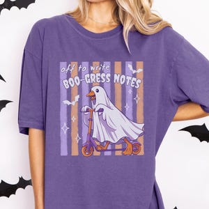 May include: A purple short-sleeve t-shirt featuring a graphic of a goose wearing a white ghost costume while riding a purple scooter. The text reads "off to write BOO-GRESS NOTES" above the goose, set against a background of vertical stripes.