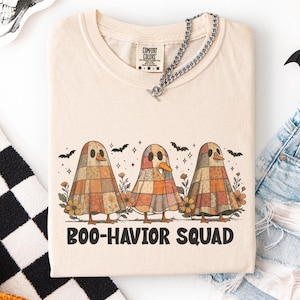 May include: Ivory-coloured t-shirt with a graphic of three cartoon ghosts in patchwork patterns, and the text "Boo-havior Squad". The design includes bats and flowers. The shirt has a crew neck and a silver chain.