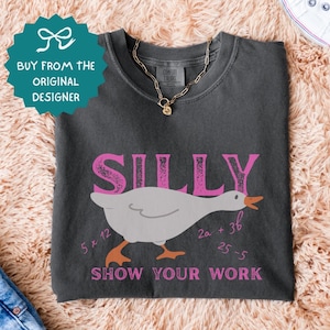 May include: Dark gray t-shirt with the word "SILLY" in pink, a cartoon goose, and the phrase "SHOW YOUR WORK" in pink. The shirt also features math equations. A gold necklace and white sneakers are visible.