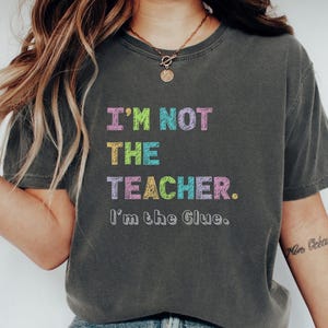 May include: A dark grey t-shirt featuring the text "I'M NOT THE TEACHER. I'm the Glue." in colourful, playful lettering. The shirt has a relaxed fit, suitable for casual wear. The shirt is a dark grey colour.