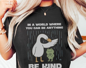 May include: Black t-shirt with the text "IN A WORLD WHERE YOU CAN BE ANYTHING BE KIND". The graphic features a cartoon goose and frog under rain. The shirt is made of a soft material.