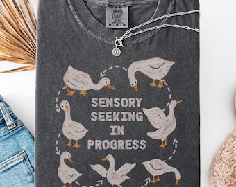 May include: A dark gray t-shirt with a circular design of geese and the text "SENSORY SEEKING IN PROGRESS". The shirt is styled with a silver necklace, blue jeans, and a white shoe. The shirt is labeled "PEPPER".