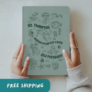 May include: A sage green journal with an aquatic theme. The cover features illustrations of sea creatures and the text "Ms. Thompson, helping progress ripple into possibility." The journal is held by hands.