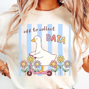 May include: An ivory t-shirt featuring a graphic of a white goose riding a pink scooter. The design includes light blue vertical stripes, colorful flowers with smiling faces, and the text "off to collect DATA" in brown and multicolored letters.