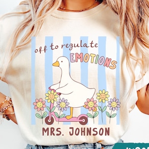 May include: Ivory-colored t-shirt with a whimsical design. A white goose rides a pink scooter amidst smiling flowers, with the text "off to regulate EMOTIONS" and "MRS. JOHNSON" in a playful font. Blue vertical stripes form the background.