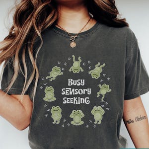 May include: Dark gray t-shirt with the text "Busy Sensory Seeking" surrounded by cartoon frogs in various poses. The shirt is a comfortable, casual style, perfect for everyday wear.