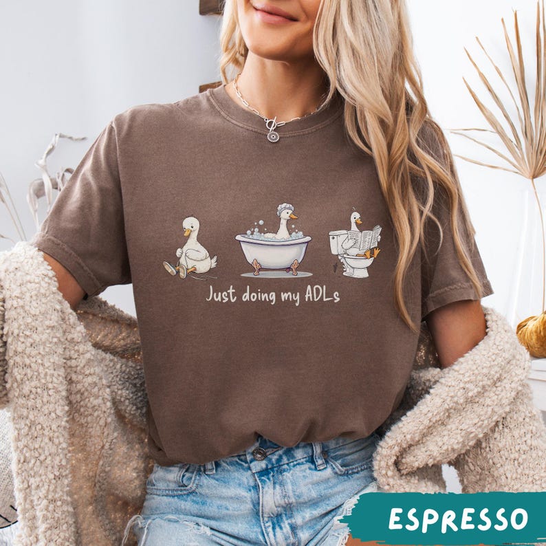 Op de afbeelding: Espresso-kleurig T-shirt met een grappige afbeelding van eenden die dagelijkse taken uitvoeren. De tekst "Just doing my ADLs" staat eronder. Het shirt is gemaakt van een zacht materiaal.