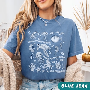May include: Blue jean colored t-shirt with a white graphic of various sea creatures and the phrases "applied behavior analysis", "helping progress ripple", and "into possibility".