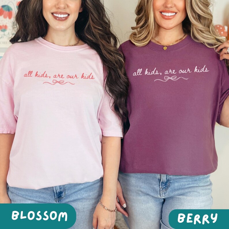 May include: Two t-shirts, one blossom pink and one berry-colored, each with the text "all kids, are our kids" in cursive script. The shirts have a relaxed fit and are paired with blue jeans. The bottom of the image has the color names.
