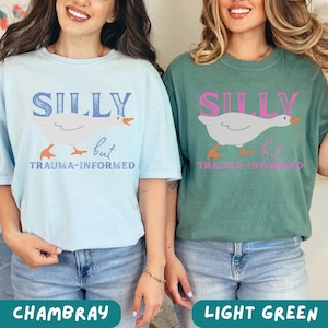 May include: Two t-shirts, one chambray blue and one light green, each with a graphic of a goose and the words "SILLY but TRAUMA-INFORMED". The goose is white with orange feet and beak. The text "SILLY" is in blue on the chambray shirt and pink on the light green shirt.