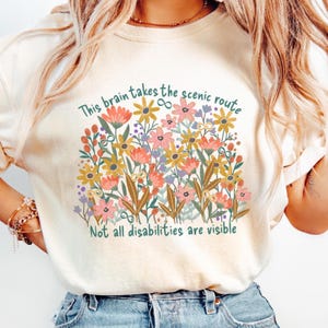 May include: Cream-coloured t-shirt with a floral design and text. The colourful floral arrangement includes pink, yellow, and orange flowers. The text reads, "This brain takes the scenic route" and "Not all disabilities are visible."