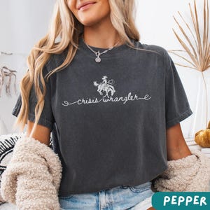 K&ouml;nnte beinhalten: Ein dunkelgraues T-Shirt mit einer wei&szlig;en Grafik einer Frau, die ein Pferd reitet, und dem Text "crisis wrangler".